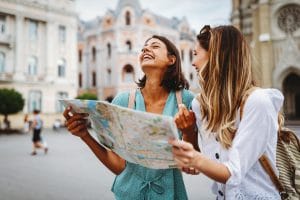 Two young women wearing short-sleeved shirts and backpacks stand in front of large, ornate buildings and laugh together, holding a partially folded paper map between them.