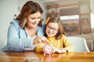 At a wooden table scattered with coins, a woman in a pale blue overshirt drops a silver coin into a pink piggy bank and smiles at the young girl in all yellow who is sitting beside her watching, also smiling.