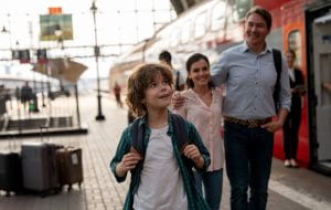 At a sunny train station, a young boy wearing a white t-shirt, green checkered overshirt, and backpack looks at a red train in wonder as a man and woman with their arms around each other watch him and smile.