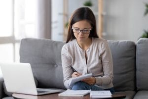 A young woman wearing black and white glasses, a gray long-sleeved shirt, and blue jeans sits on a plush gray couch and writes on a spiral notepad intently, a laptop and two more notebooks resting on the small table in front of her.