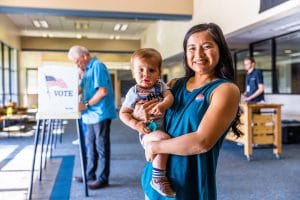 A smiling woman holding a baby stands in a voting center, both wearing "I Voted" stickers. In the background, an older man is casting his vote at a booth with a "VOTE" sign featuring an American flag.